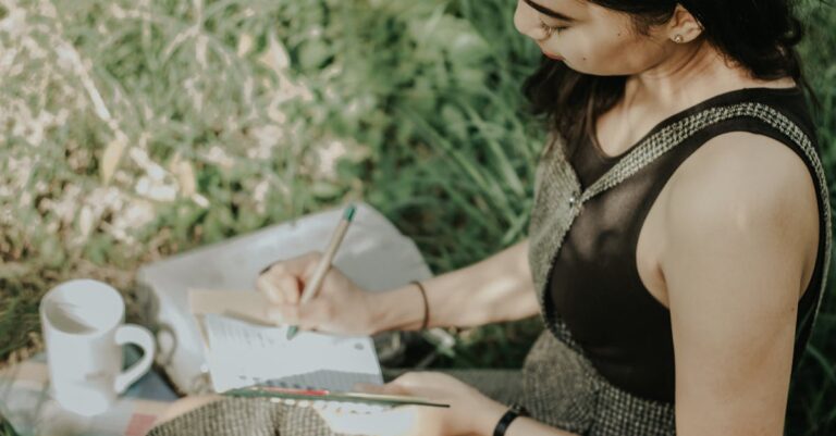 Uma mulher sentada na grama, escrevendo em um caderno, com uma caneta e uma xícara de café ao lado.